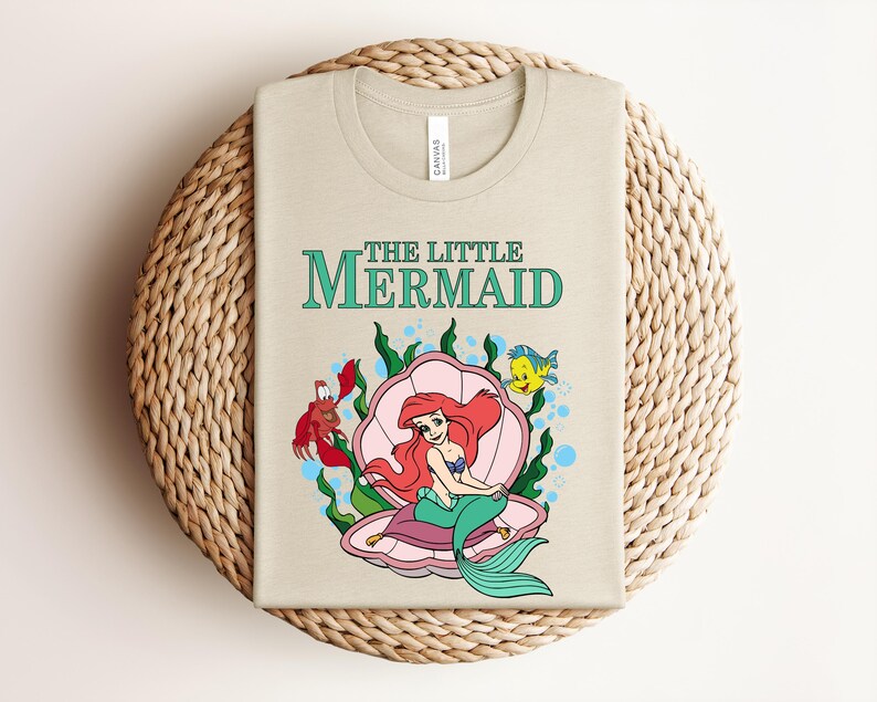 May include: A beige crew neck t-shirt featuring a graphic of The Little Mermaid with Ariel sitting in a pink seashell, accompanied by a red crab and a yellow and blue fish, surrounded by green seaweed and blue bubbles on the front of the shirt.