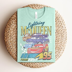 Puede incluir: Una camiseta verde claro con un gr&aacute;fico de Rayo McQueen y otros personajes de Cars. El dise&ntilde;o incluye el texto "Lightning McQueen" y "95", con la frase "Think Fast" en la parte inferior. La camiseta est&aacute; doblada sobre una estera tejida.