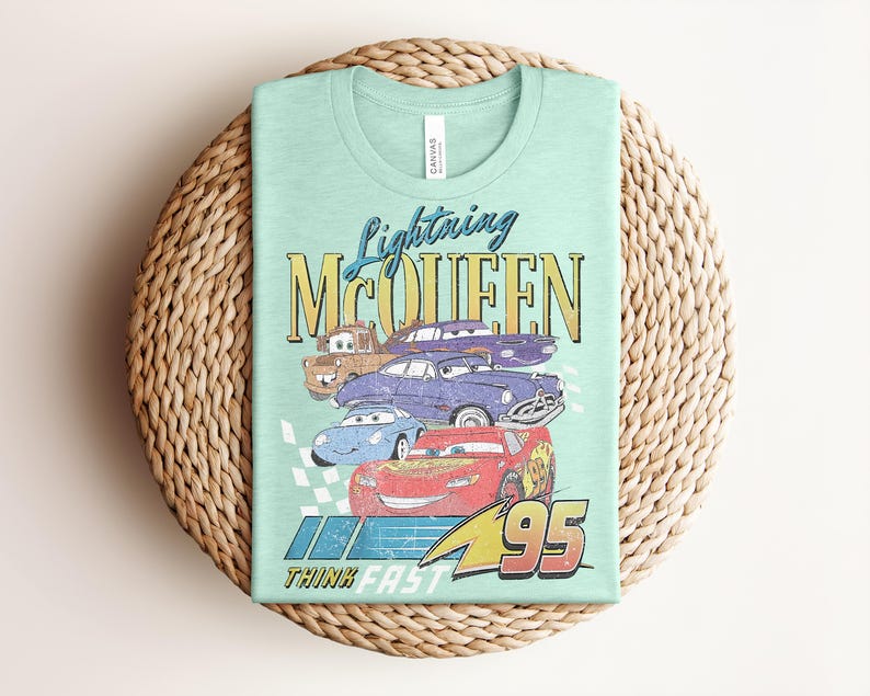 Puede incluir: Una camiseta verde claro con un gr&aacute;fico de Rayo McQueen y otros personajes de Cars. El dise&ntilde;o incluye el texto "Lightning McQueen" y "95", con la frase "Think Fast" en la parte inferior. La camiseta est&aacute; doblada sobre una estera tejida.