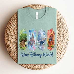 May include: A light green t-shirt featuring watercolor illustrations of iconic Walt Disney World landmarks. The design includes the Tree of Life, Cinderella Castle, Spaceship Earth, and the Tower of Terror, with "Walt Disney World" written below.