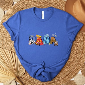 K&ouml;nnte beinhalten: Ein blaues T-Shirt mit dem Wort "NANA" in bunten, Auto-Themen-Buchstaben. Das Design umfasst Rennwagen und Zielflaggen. Das Shirt besteht aus einem weichen Material und ist ein tolles Geschenk.