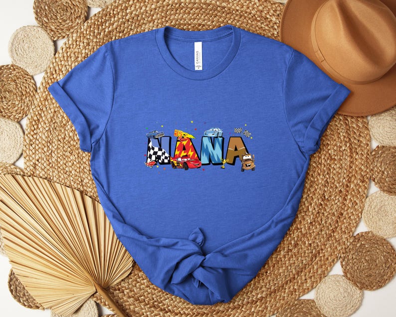 K&ouml;nnte beinhalten: Ein blaues T-Shirt mit dem Wort "NANA" in bunten, Auto-Themen-Buchstaben. Das Design umfasst Rennwagen und Zielflaggen. Das Shirt besteht aus einem weichen Material und ist ein tolles Geschenk.