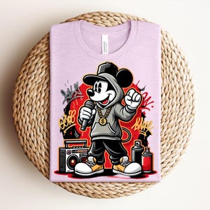 May include: A light purple t-shirt featuring a cartoon Mickey Mouse in a hip-hop style. Mickey is wearing a gray hoodie, black pants, and a gold chain, holding a microphone. Graffiti art and a boombox are also visible.