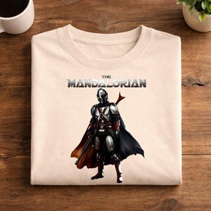 Op de afbeelding: Cr&egrave;mekleurige crewneck sweatshirt met een afbeelding van The Mandalorian. De afbeelding toont het personage in volledige bepantsering met een zwarte cape en een blaster. De tekst "THE MANDALORIAN" is in een metallic lettertype boven het personage gedrukt.