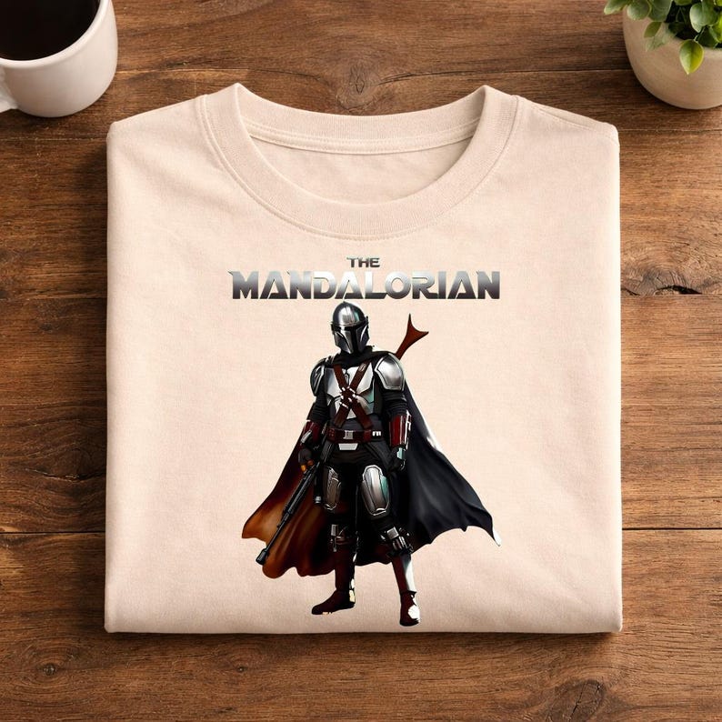 Op de afbeelding: Cr&egrave;mekleurige crewneck sweatshirt met een afbeelding van The Mandalorian. De afbeelding toont het personage in volledige bepantsering met een zwarte cape en een blaster. De tekst "THE MANDALORIAN" is in een metallic lettertype boven het personage gedrukt.