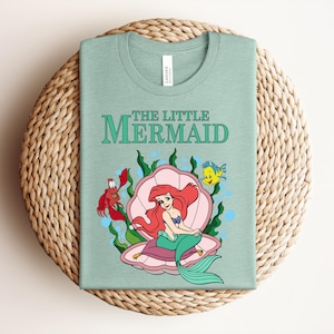May include: A light green t-shirt featuring a graphic of The Little Mermaid with Ariel sitting in a pink seashell, accompanied by a red crab and a yellow fish, surrounded by green seaweed and blue bubbles, with the text The Little Mermaid above.