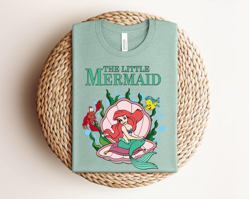 May include: A light green t-shirt featuring a graphic of The Little Mermaid with Ariel sitting in a pink seashell, accompanied by a red crab and a yellow fish, surrounded by green seaweed and blue bubbles, with the text The Little Mermaid above.