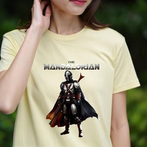 Op de afbeelding: Een lichtgeel t-shirt met de tekst "THE MANDALORIAN" boven een afbeelding van het Mandalorian-personage in volledige uitrusting, cape en wapen. Het ontwerp is gecentreerd op het shirt.