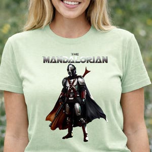 Op de afbeelding: Lichtgroen T-shirt met de tekst "THE MANDALORIAN" boven een illustratie van het Mandalorian-personage in zilveren harnas, een zwarte cape en een blaster. Het ontwerp is gecentreerd op het shirt.
