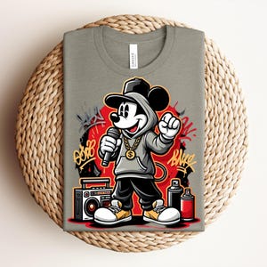 May include: A light olive green t-shirt featuring a cartoon Mickey Mouse character in a hip-hop style. Mickey is wearing a gray hoodie, gold chain, and holding a microphone. Graffiti art and a boombox are also featured in the design.