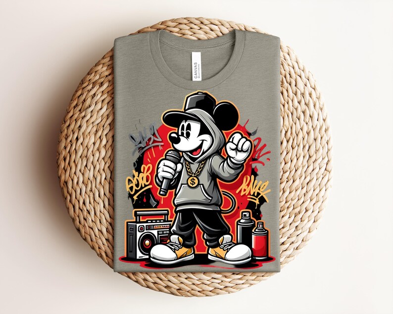 May include: A light olive green t-shirt featuring a cartoon Mickey Mouse character in a hip-hop style. Mickey is wearing a gray hoodie, gold chain, and holding a microphone. Graffiti art and a boombox are also featured in the design.