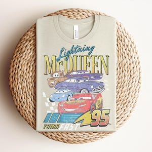Puede incluir: Una camiseta verde claro con un gr&aacute;fico de Rayo McQueen y otros personajes de Cars. El dise&ntilde;o incluye el texto "Lightning McQueen" y "95", con la frase "Think Fast" debajo. La camiseta est&aacute; doblada sobre una superficie tejida.