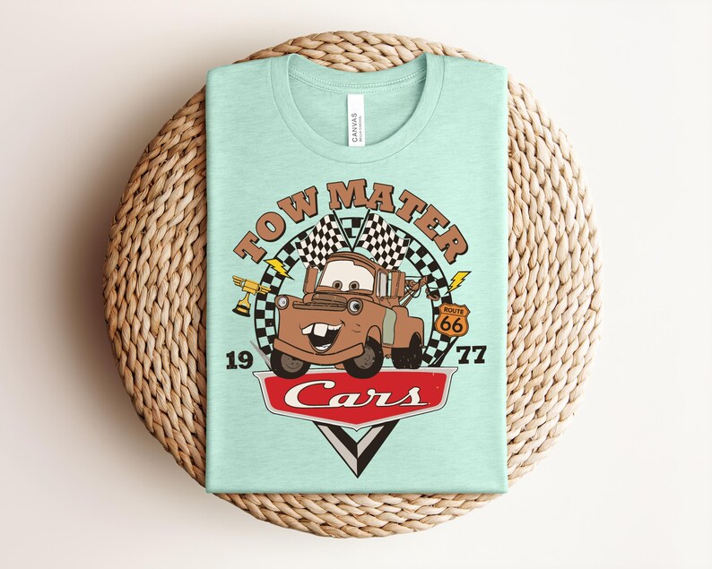 Puede incluir: Una camiseta de color verde menta claro con un gr&aacute;fico del personaje Tow Mater de la pel&iacute;cula Cars. El dise&ntilde;o incluye el texto "TOW MATER", "1977", "Route 66" y "Cars" con banderas a cuadros, un trofeo y rayos como detalles gr&aacute;ficos.