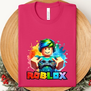 May include: A magenta t-shirt featuring a colorful graphic of a block-style character with headphones holding a game controller. The word "ROBLOX" is printed in rainbow colors below the character. The shirt is folded on a wooden surface.