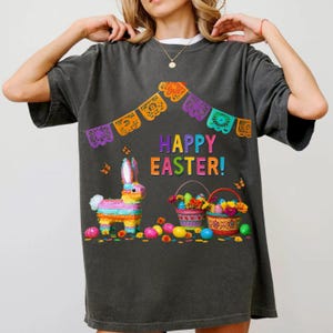 K&ouml;nnte beinhalten: Dunkelgraues T-Shirt mit einem farbenfrohen "Frohe Ostern!"-Motiv. Das Design umfasst eine Pi&ntilde;ata, Osterk&ouml;rbe, Schmetterlinge und dekorative Banner. Das Shirt hat eine lockere Passform.