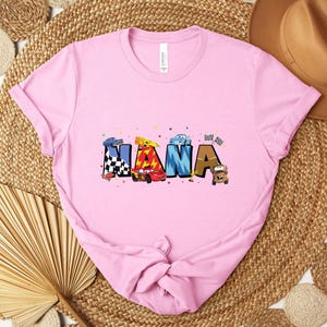 K&ouml;nnte beinhalten: Hellrosa T-Shirt mit dem Wort "NANA" in bunten Buchstaben im Auto-Design. Die Buchstaben sind mit Rennwagen und Zielflaggen verziert. Das T-Shirt ist aus einem weichen Material.