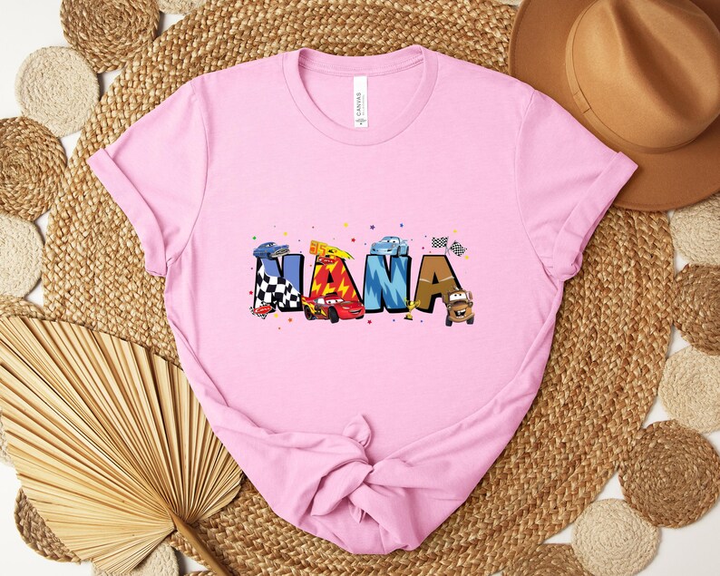 K&ouml;nnte beinhalten: Hellrosa T-Shirt mit dem Wort "NANA" in bunten Buchstaben im Auto-Design. Die Buchstaben sind mit Rennwagen und Zielflaggen verziert. Das T-Shirt ist aus einem weichen Material.