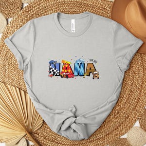 K&ouml;nnte beinhalten: Hellgraues T-Shirt mit dem Wort "NANA" in bunten Buchstaben im Autothema. Die Buchstaben sind mit Rennwagen, Schachflaggen und anderen Rennsportelementen verziert. Das Shirt ist aus weichem Material.
