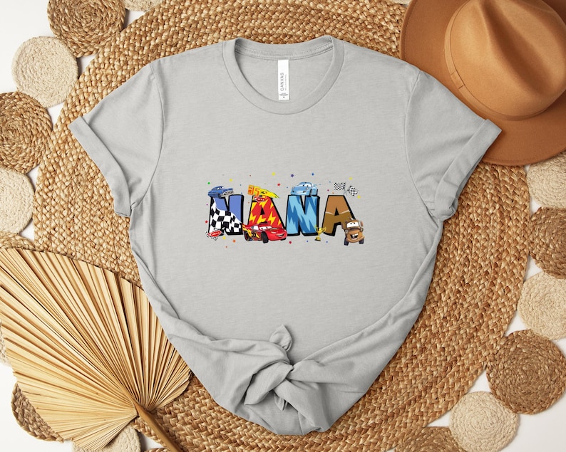K&ouml;nnte beinhalten: Hellgraues T-Shirt mit dem Wort "NANA" in bunten Buchstaben im Autothema. Die Buchstaben sind mit Rennwagen, Schachflaggen und anderen Rennsportelementen verziert. Das Shirt ist aus weichem Material.