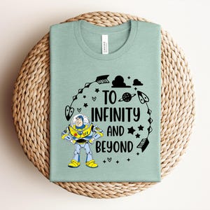 Peut inclure: Un t-shirt vert clair avec le texte "TO INFINITY AND BEYOND" et un dessin anim&eacute; de Buzz l'&Eacute;clair. Le motif comprend des &eacute;toiles, des plan&egrave;tes et des fl&egrave;ches. Le t-shirt est pli&eacute; sur un tapis tiss&eacute;.