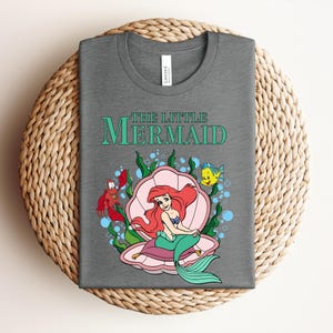 May include: A heather gray t-shirt featuring the text THE LITTLE MERMAID in teal. The graphic shows a red-haired mermaid with a green tail sitting inside a pink seashell, accompanied by a red crab and a yellow fish amidst green seaweed and blue bubbles.