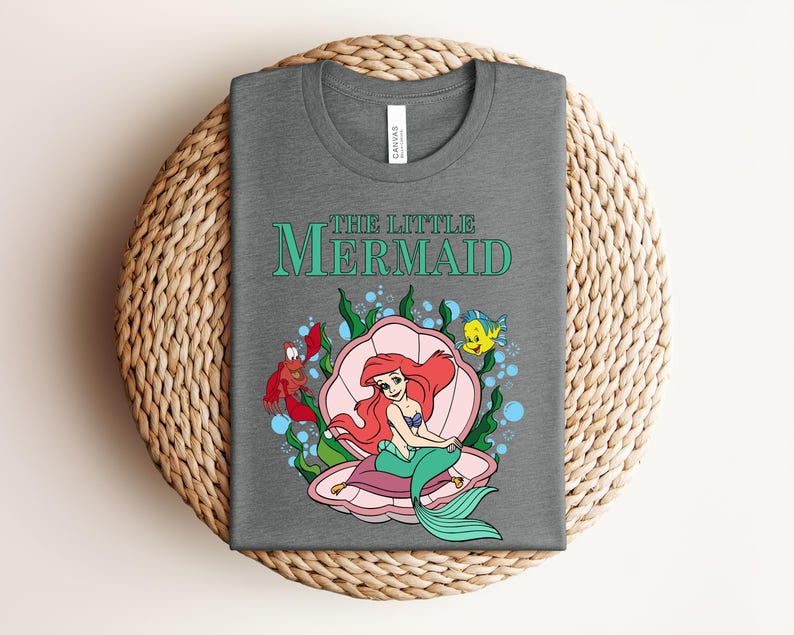 May include: A heather gray t-shirt featuring the text THE LITTLE MERMAID in teal. The graphic shows a red-haired mermaid with a green tail sitting inside a pink seashell, accompanied by a red crab and a yellow fish amidst green seaweed and blue bubbles.