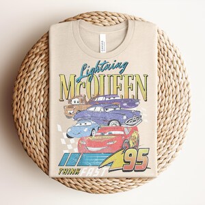 Puede incluir: Camiseta beige con un gr&aacute;fico de Rayo McQueen y otros personajes de Cars. El dise&ntilde;o incluye el texto "Lightning McQueen", "95" y "Think Fast". La camiseta est&aacute; doblada sobre una superficie circular tejida.