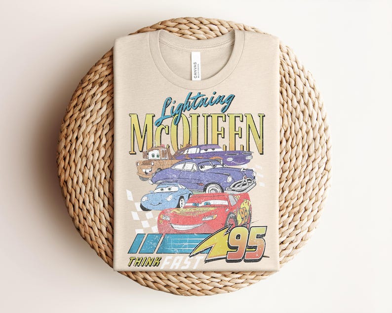 Puede incluir: Camiseta beige con un gr&aacute;fico de Rayo McQueen y otros personajes de Cars. El dise&ntilde;o incluye el texto "Lightning McQueen", "95" y "Think Fast". La camiseta est&aacute; doblada sobre una superficie circular tejida.