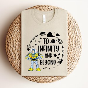 Peut inclure: Un t-shirt beige avec une illustration de dessin anim&eacute; de Buzz l'&Eacute;clair et l'inscription "To Infinity and Beyond". Le motif comprend des &eacute;toiles, des plan&egrave;tes et des fl&egrave;ches noires. Le t-shirt est pr&eacute;sent&eacute; sur une surface circulaire tiss&eacute;e.
