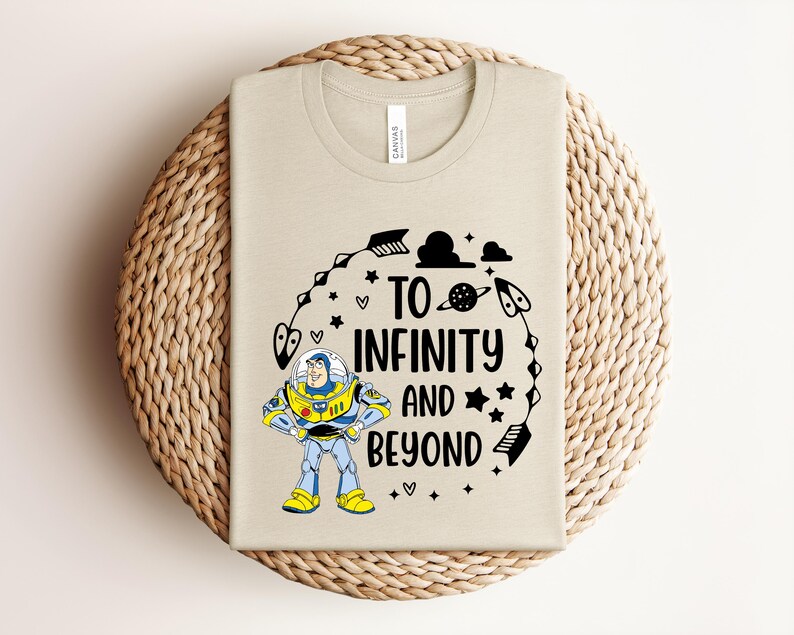 Peut inclure: Un t-shirt beige avec une illustration de dessin anim&eacute; de Buzz l'&Eacute;clair et l'inscription "To Infinity and Beyond". Le motif comprend des &eacute;toiles, des plan&egrave;tes et des fl&egrave;ches noires. Le t-shirt est pr&eacute;sent&eacute; sur une surface circulaire tiss&eacute;e.