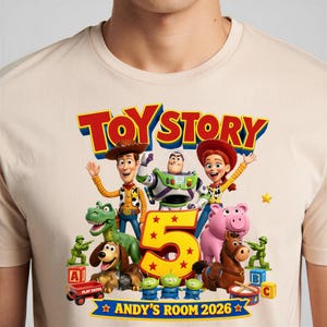 May include: A beige t-shirt featuring a colorful Toy Story graphic with Woody, Buzz Lightyear, Jessie, Rex, Slinky Dog, Bullseye, and three aliens. The design includes a large yellow number 5 and the text "TOY STORY" and "ANDY'S ROOM 2026".