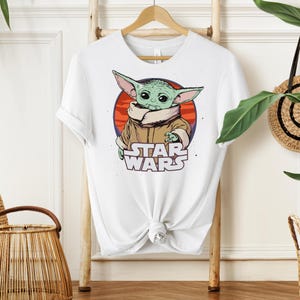 Puede incluir: Camiseta blanca con un dise&ntilde;o de Grogu, tambi&eacute;n conocido como Baby Yoda, de Star Wars. El dise&ntilde;o muestra a Grogu dentro de un c&iacute;rculo rojo y naranja, con las palabras "STAR WARS" debajo.