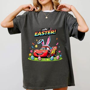 Puede incluir: Camiseta gris oscuro con un coche de carreras rojo de dibujos animados con orejas de conejo, rodeado de huevos de Pascua y flores. El texto "HAPPY EASTER!" est&aacute; encima del coche. Un dise&ntilde;o festivo para la Pascua.