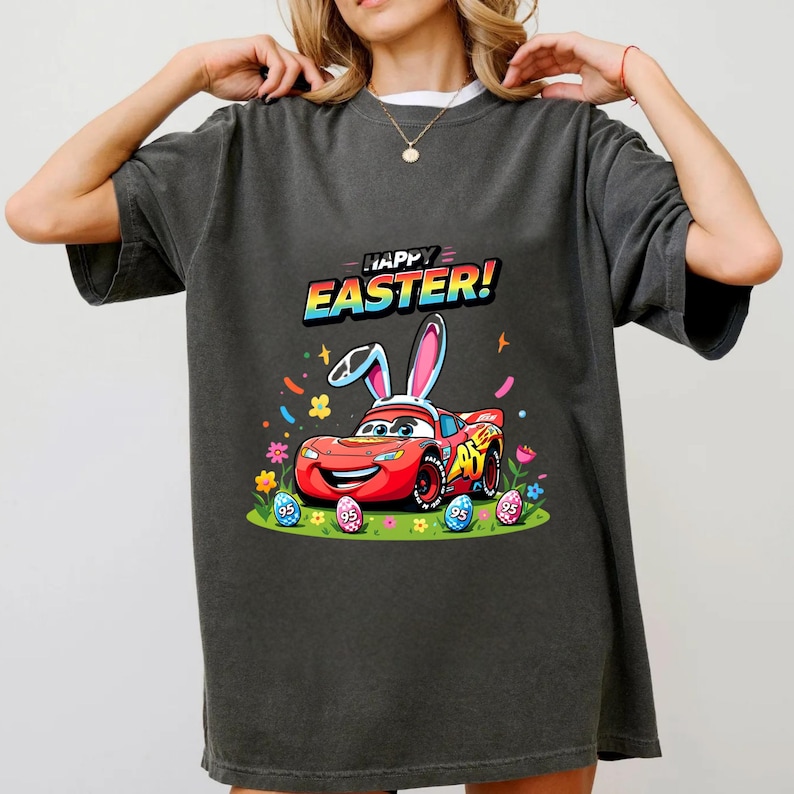 Puede incluir: Camiseta gris oscuro con un coche de carreras rojo de dibujos animados con orejas de conejo, rodeado de huevos de Pascua y flores. El texto "HAPPY EASTER!" est&aacute; encima del coche. Un dise&ntilde;o festivo para la Pascua.