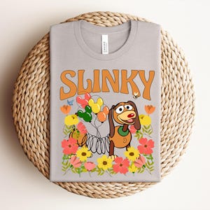 K&ouml;nnte beinhalten: Ein hellgraues T-Shirt mit dem Wort "SLINKY" in Orange. Das Shirt hat eine Grafik von Slinky Dog aus Toy Story, umgeben von bunten Blumen, Luftballons und einer Biene.