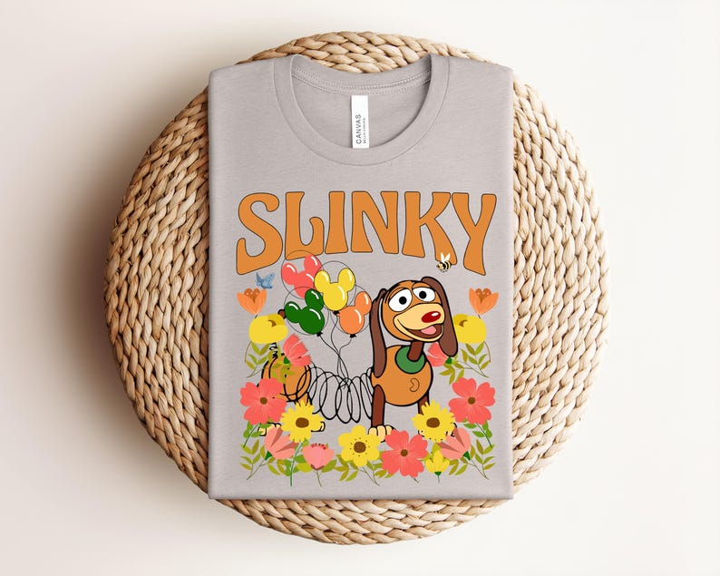 K&ouml;nnte beinhalten: Ein hellgraues T-Shirt mit dem Wort "SLINKY" in Orange. Das Shirt hat eine Grafik von Slinky Dog aus Toy Story, umgeben von bunten Blumen, Luftballons und einer Biene.