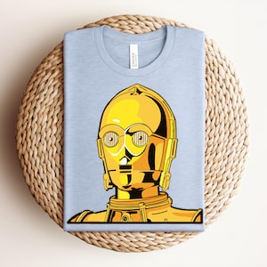 May include: A light blue t-shirt featuring a gold and yellow graphic of the Star Wars character C-3PO. The graphic is a detailed illustration of the droid's head and upper body. The shirt is folded and placed on a woven, circular surface.