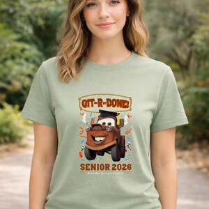 Op de afbeelding: Saliegroen T-shirt met een cartoonafbeelding van Takel uit de film Cars, met een afstudeerhoed. De tekst "GIT-R-DONE!" staat boven Takel en "SENIOR 2026" eronder. Het shirt heeft een ronde hals.