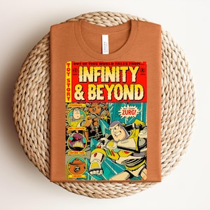 May include: A rust-colored t-shirt with a comic book-style graphic. The graphic features the words "INFINITY & BEYOND" in large letters, along with illustrations of characters and the text "TOY STORY". The shirt is folded on a woven surface.