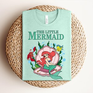 May include: A light mint green t-shirt featuring a graphic print of The Little Mermaid with Ariel sitting in a pink seashell, accompanied by a red crab and a yellow fish, surrounded by green seaweed and blue bubbles on the front of the garment.