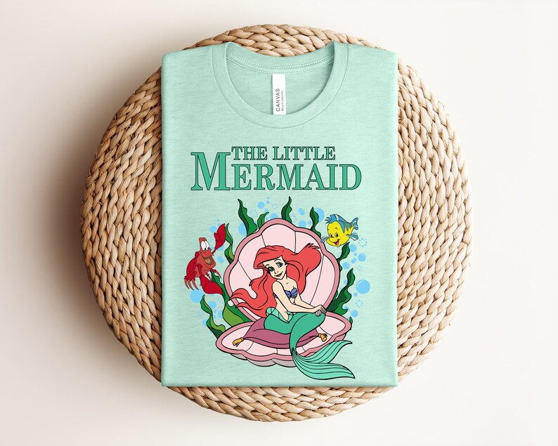 May include: A light mint green t-shirt featuring a graphic print of The Little Mermaid with Ariel sitting in a pink seashell, accompanied by a red crab and a yellow fish, surrounded by green seaweed and blue bubbles on the front of the garment.