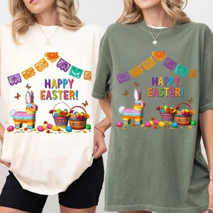 K&ouml;nnte beinhalten: Cremefarbene und olivgr&uuml;ne T-Shirts mit einem farbenfrohen "Frohe Ostern!"-Motiv. Das Design umfasst einen Pi&ntilde;ata-Hasen, Osterk&ouml;rbe, Schmetterlinge und dekorative Papierbanner. Die Shirts sind l&auml;ssig und festlich.
