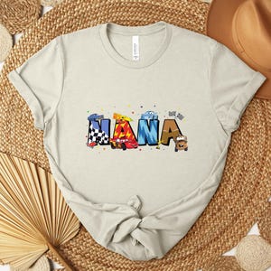 K&ouml;nnte beinhalten: Ein hellbeige T-Shirt mit dem Wort "NANA" in bunten, cartoonartigen Buchstaben. Jeder Buchstabe zeigt ein Rennwagen-Design mit karierten Flaggen und Autoillustrationen. Das Shirt besteht aus weichem Material.