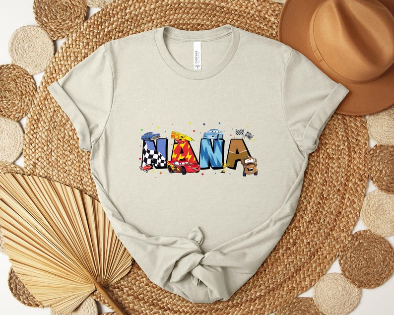 K&ouml;nnte beinhalten: Ein hellbeige T-Shirt mit dem Wort "NANA" in bunten, cartoonartigen Buchstaben. Jeder Buchstabe zeigt ein Rennwagen-Design mit karierten Flaggen und Autoillustrationen. Das Shirt besteht aus weichem Material.