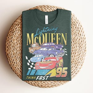 Puede incluir: Camiseta verde oscuro con un gr&aacute;fico de Rayo McQueen y otros personajes de Cars. El dise&ntilde;o incluye el texto "Lightning McQueen" y "Think Fast 95" en un estilo vintage. La camiseta est&aacute; doblada sobre una superficie tejida.