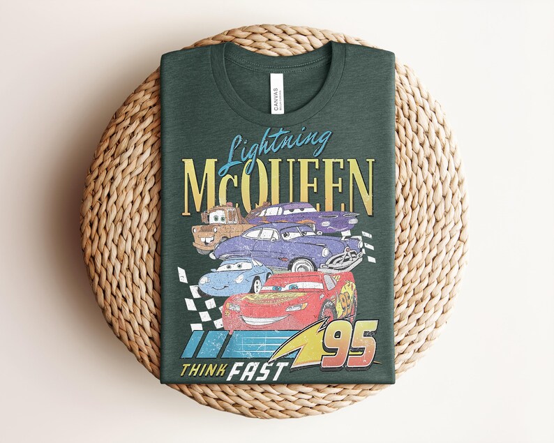 Puede incluir: Camiseta verde oscuro con un gr&aacute;fico de Rayo McQueen y otros personajes de Cars. El dise&ntilde;o incluye el texto "Lightning McQueen" y "Think Fast 95" en un estilo vintage. La camiseta est&aacute; doblada sobre una superficie tejida.