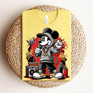 May include: Yellow t-shirt featuring a cartoon illustration of Mickey Mouse in a hip-hop style. Mickey is wearing a gray hoodie, black pants, and a gold chain, holding a microphone. The design includes graffiti art and a boombox.
