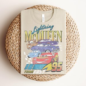 Puede incluir: Camiseta color beige con un dise&ntilde;o de Rayo McQueen y otros personajes de Cars. El dise&ntilde;o incluye el texto "Lightning McQueen" y "95", con la frase "Think Fast" en la parte inferior. La camiseta est&aacute; hecha de un material suave.
