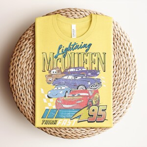 Puede incluir: Camiseta amarilla con un dise&ntilde;o de Rayo McQueen y otros personajes de la pel&iacute;cula Cars. El dise&ntilde;o incluye el texto "Lightning McQueen" y "95", con una bandera a cuadros y las palabras "Think Fast".