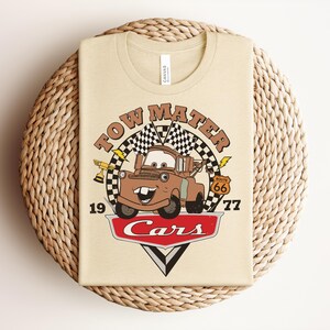 Puede incluir: Una camiseta de color crema con un gr&aacute;fico del personaje Tow Mater de la pel&iacute;cula Cars. El dise&ntilde;o incluye el texto Tow Mater, 1977, Route 66 y Cars, junto con banderas a cuadros, un trofeo y detalles de rayos.